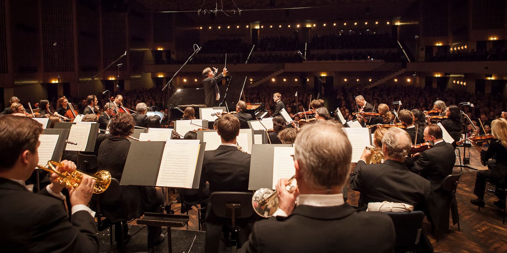 Ein Orchester spielt auf einer Bühne vor Publikum, der Dirigent steht im Zentrum und dirigiert.