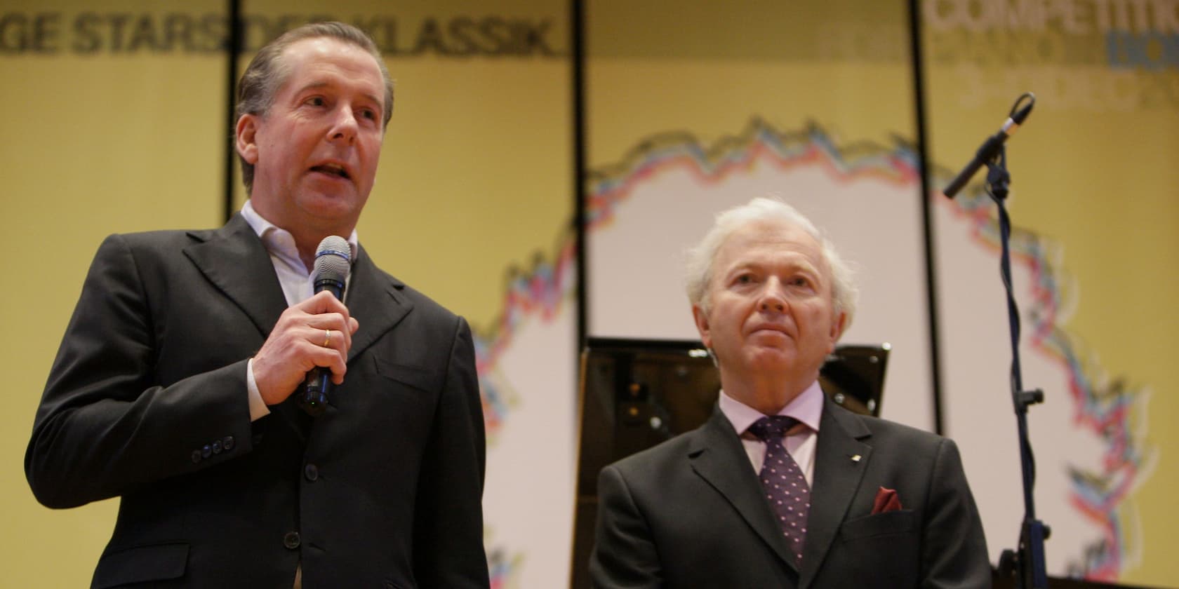 Two men standing on a stage speaking into microphones at a classical music competition.