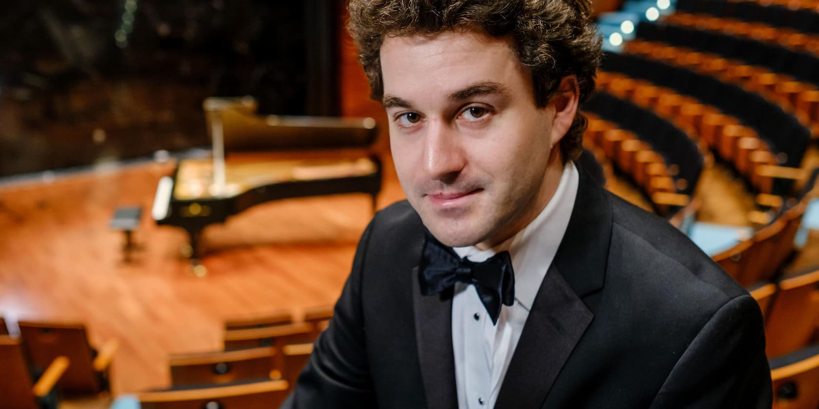 A smiling man in a suit sits in an empty concert hall with a grand piano in the background.