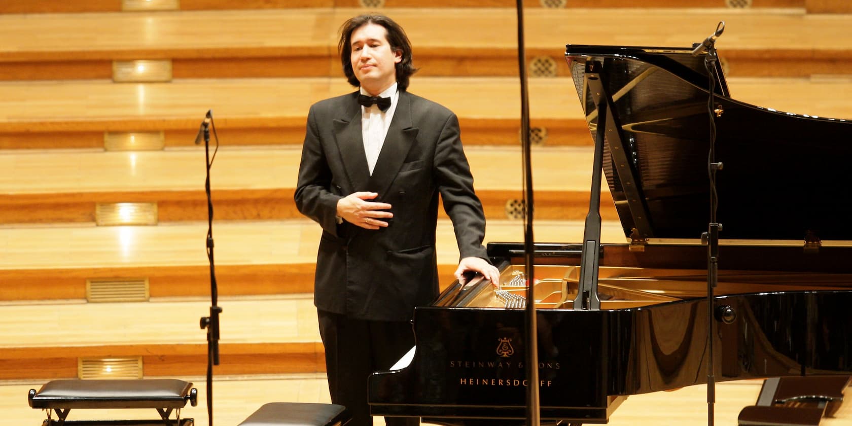 Man in a suit stands next to a grand piano in a concert hall.