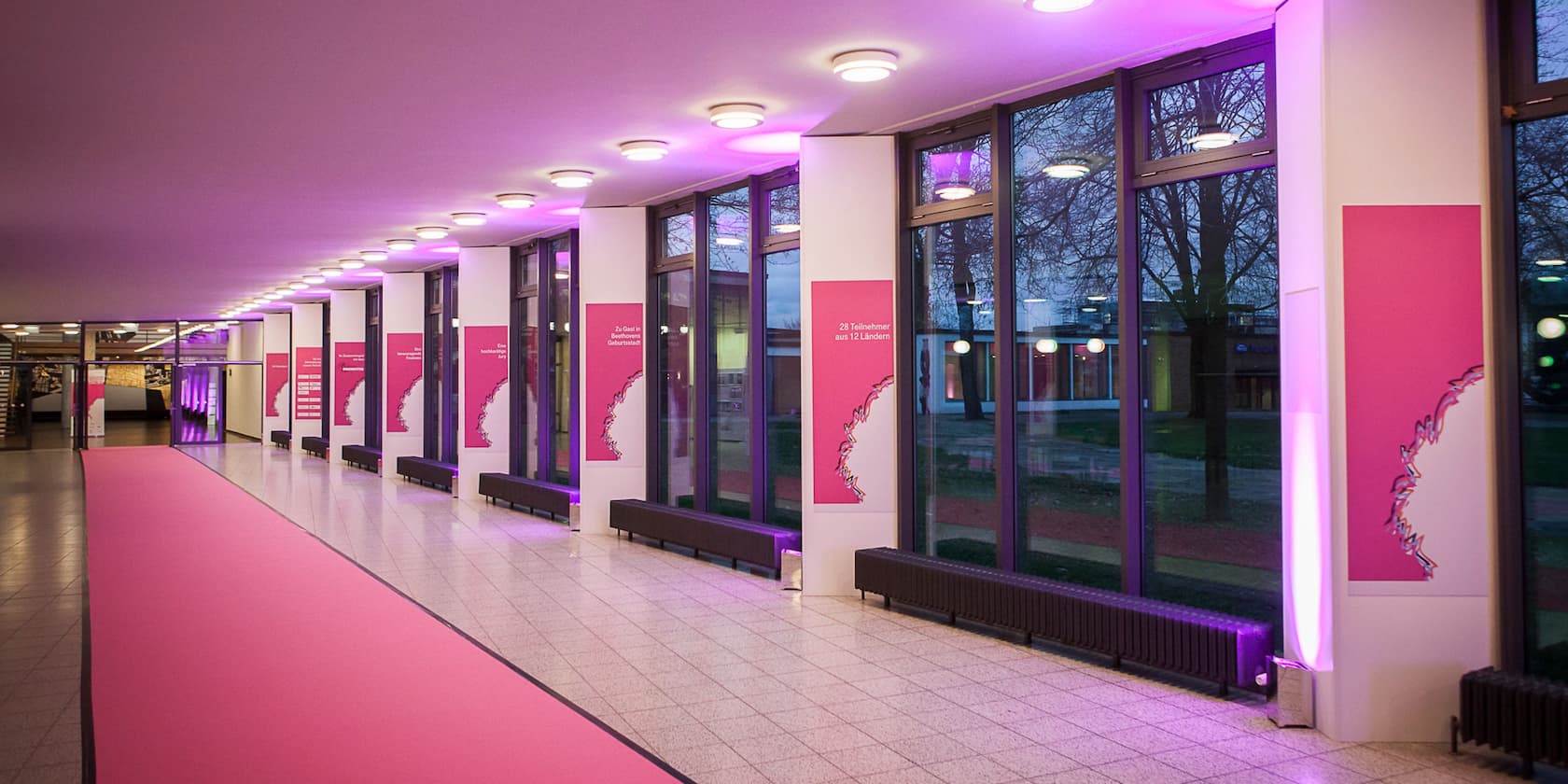 A long, brightly lit hallway with large windows and a pink carpet. Posters with text in pink frames are mounted on the walls.