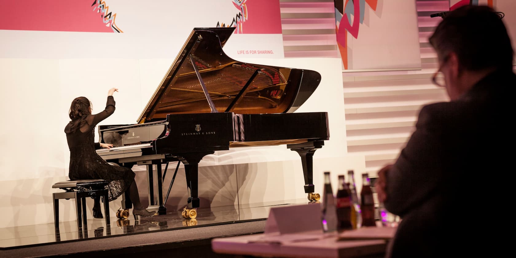 A woman is playing the piano on a stage, with a quote in the background: 'Life is for sharing'.