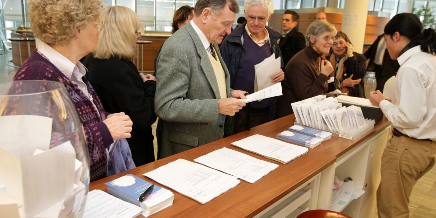 Several elderly people are standing at an information desk, looking at documents. An employee stands behind the desk.