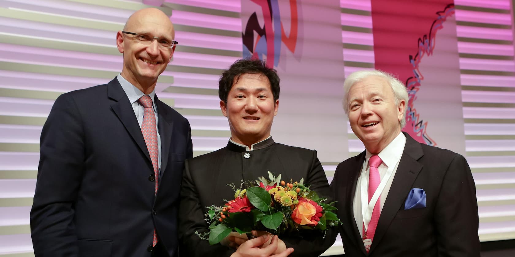 Three men in formal attire, with one in the middle holding a bouquet of flowers.