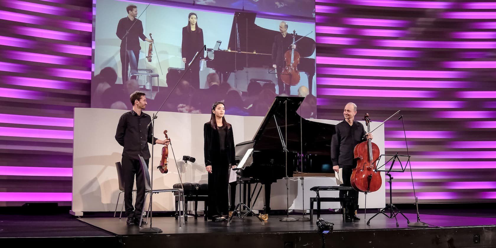 Three musicians stand on stage with a purple-lit background, in front of a piano. One holds a violin, another a cello.