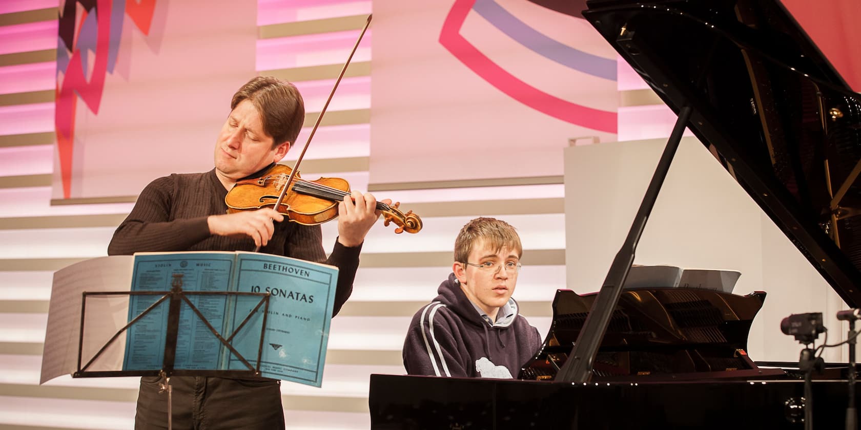 A violinist and a pianist perform a musical piece together. In front of the violinist is a music sheet with the title 'Beethoven 10 Sonatas'.