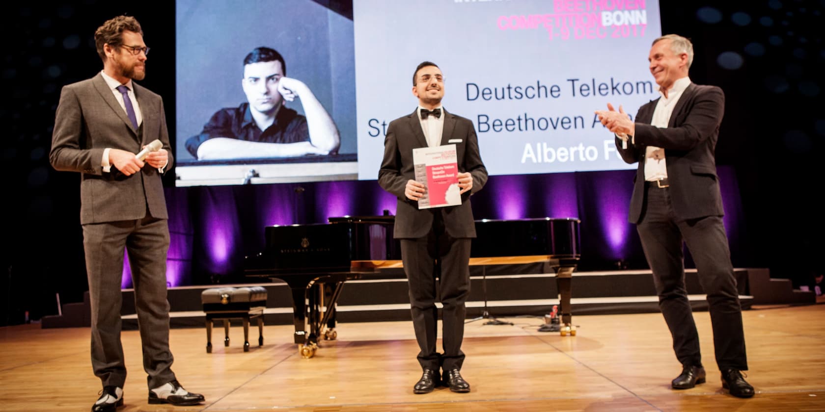 Preisverleihung beim Telekom Beethoven Wettbewerb Bonn 2017. Ein Sieger steht mit einem Zertifikat auf der Bühne zwischen zwei Moderatoren.