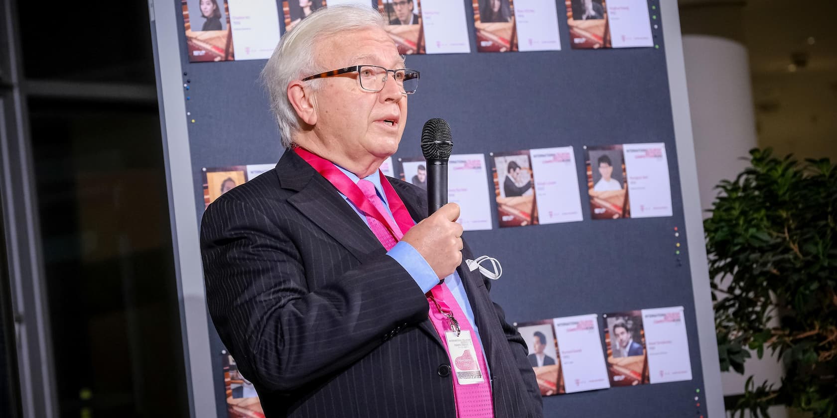 A man is speaking into a microphone at an event. In the background, several photos of people are displayed on a pinboard.