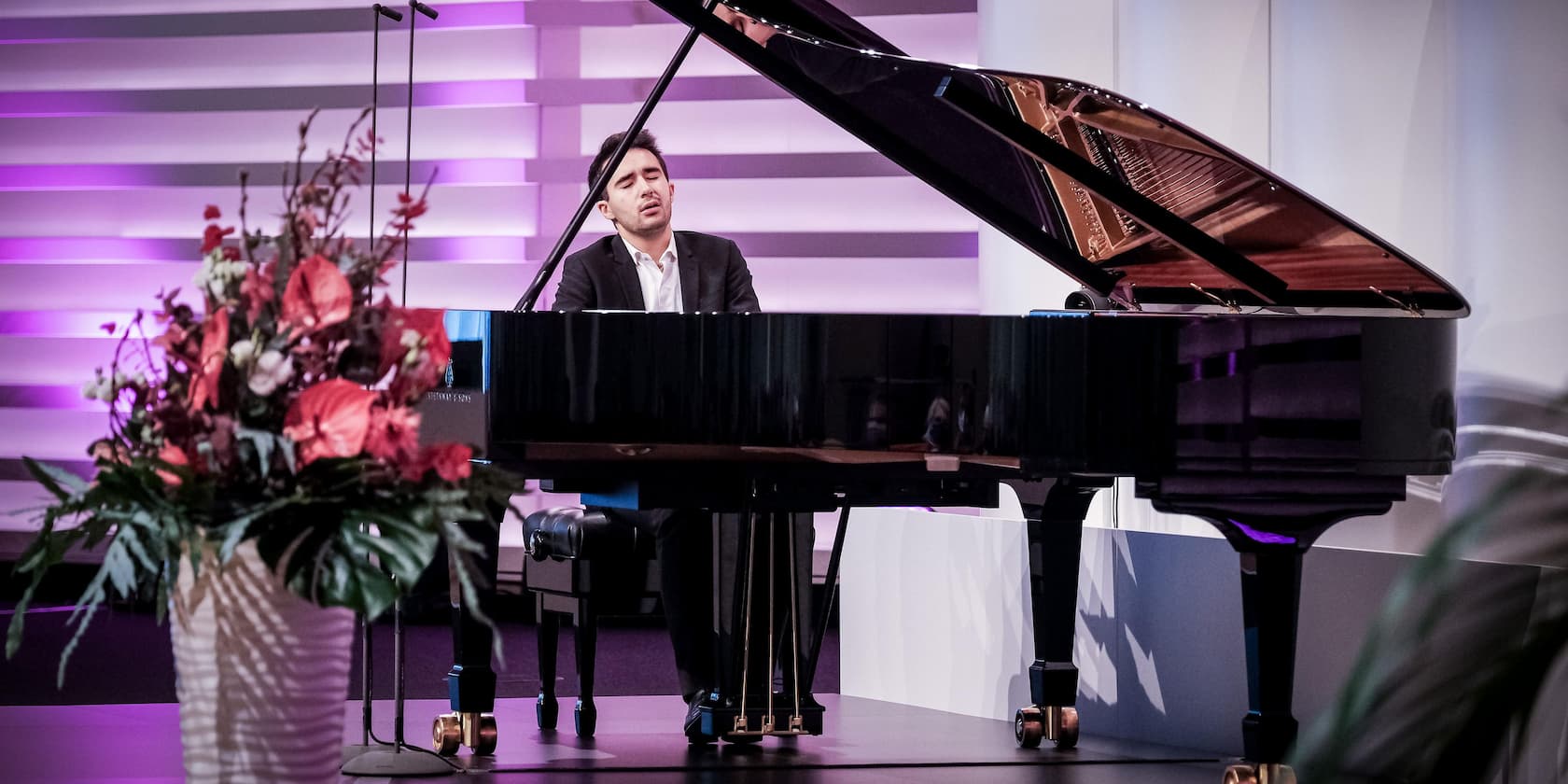 A man is playing piano on stage, with a vase of flowers in the foreground.