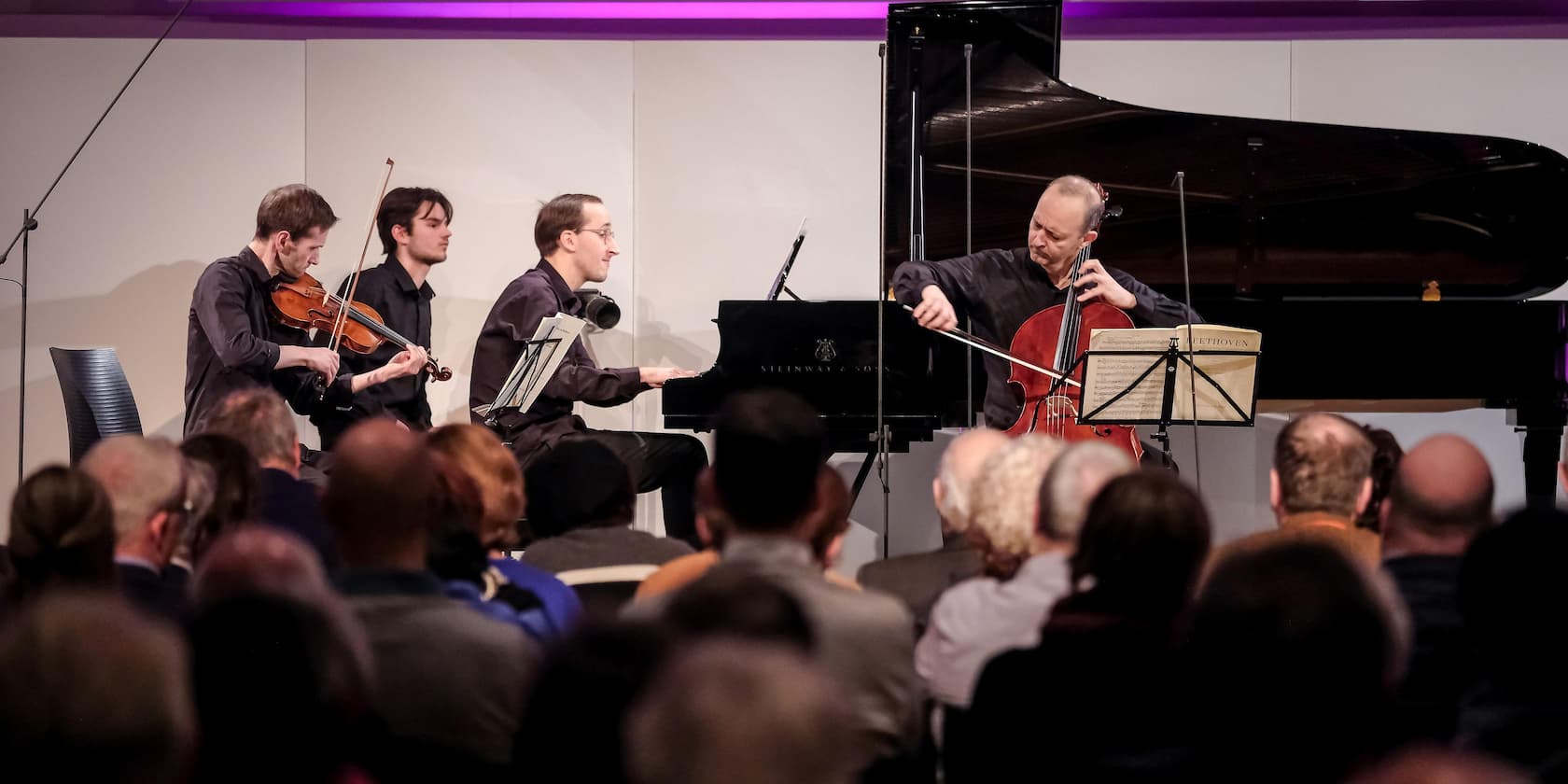 A string quartet performs before an audience in a concert hall. Two violinists, a cellist, and a pianist are visible.