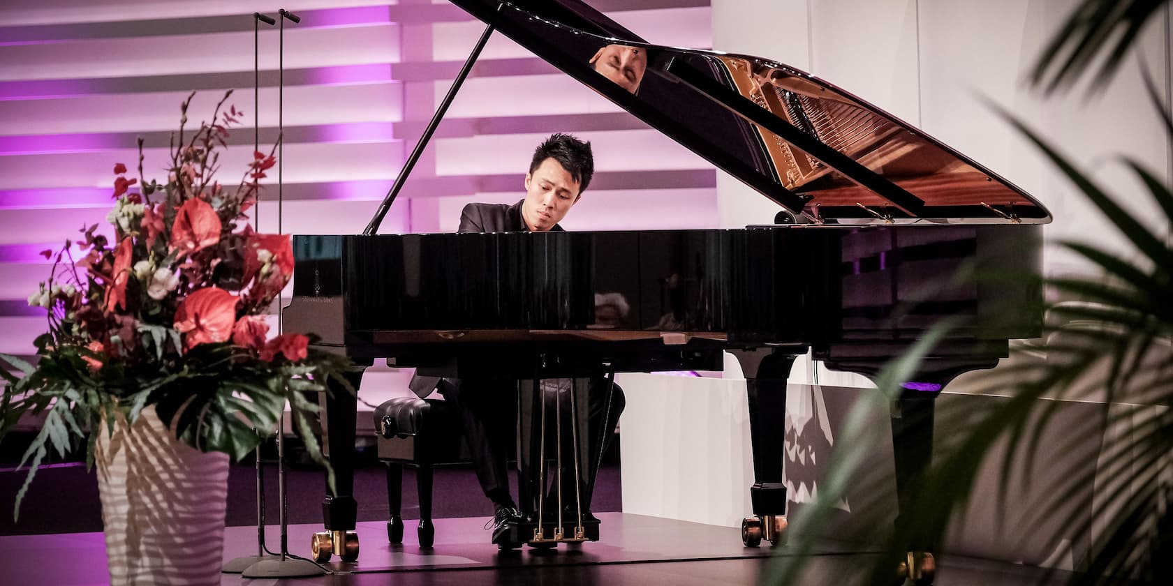 A musician plays piano on a stage in front of a wall with purple lighting. A flower arrangement is in the foreground.