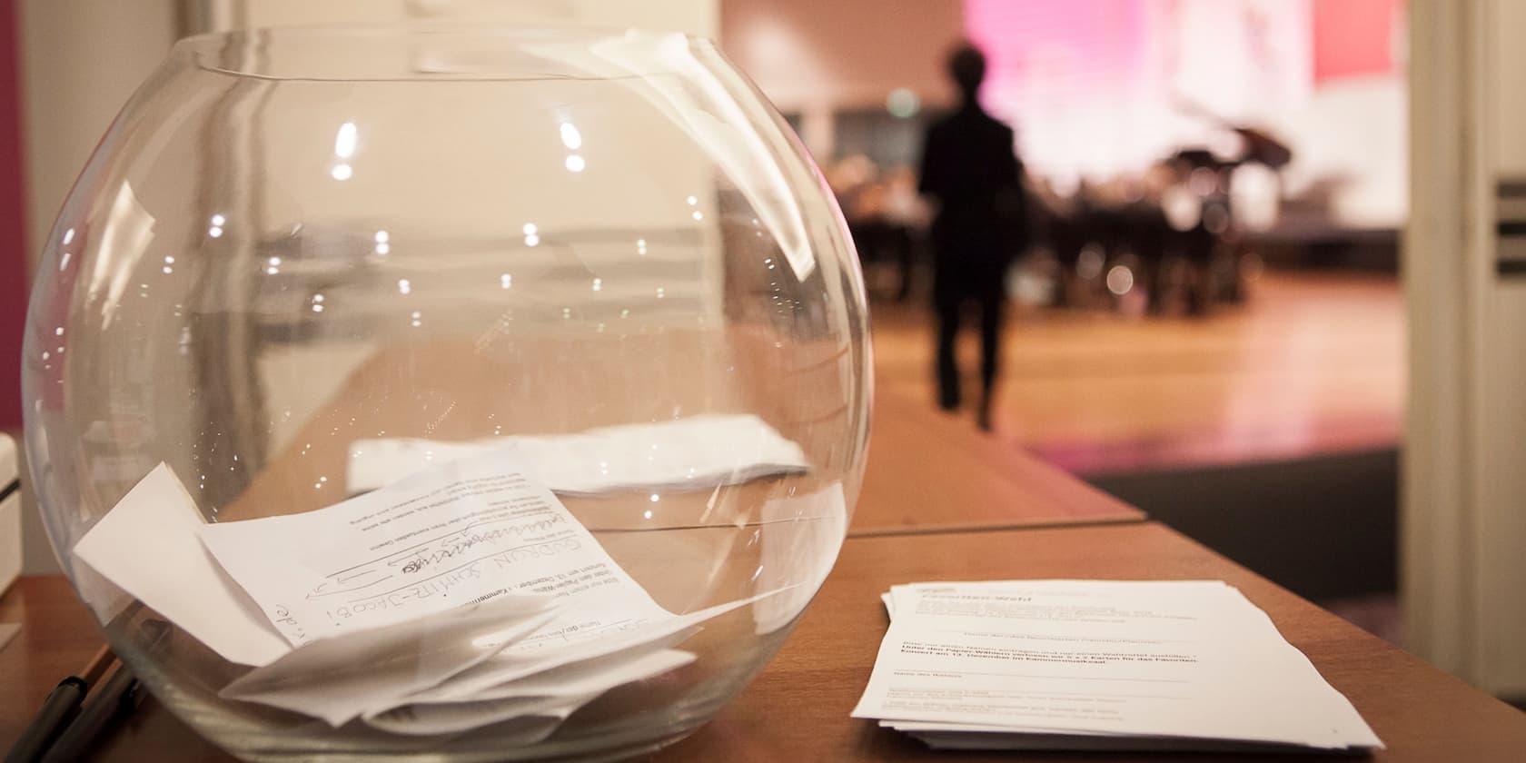 Large glass bowl with several sheets of paper inside, standing on a table. A person is blurred in the background.