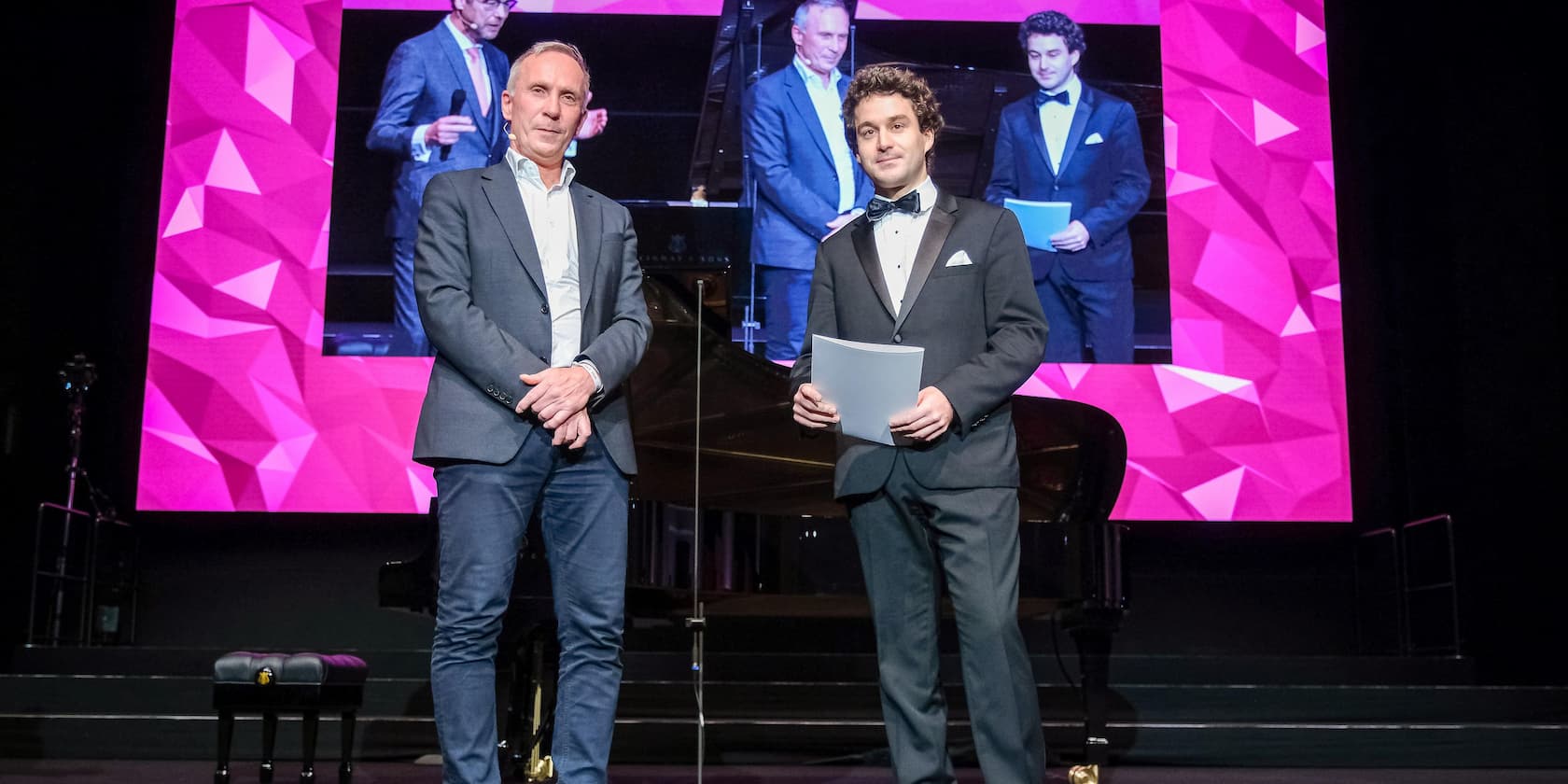 Two men standing on a stage in front of a large screen with a purple background.