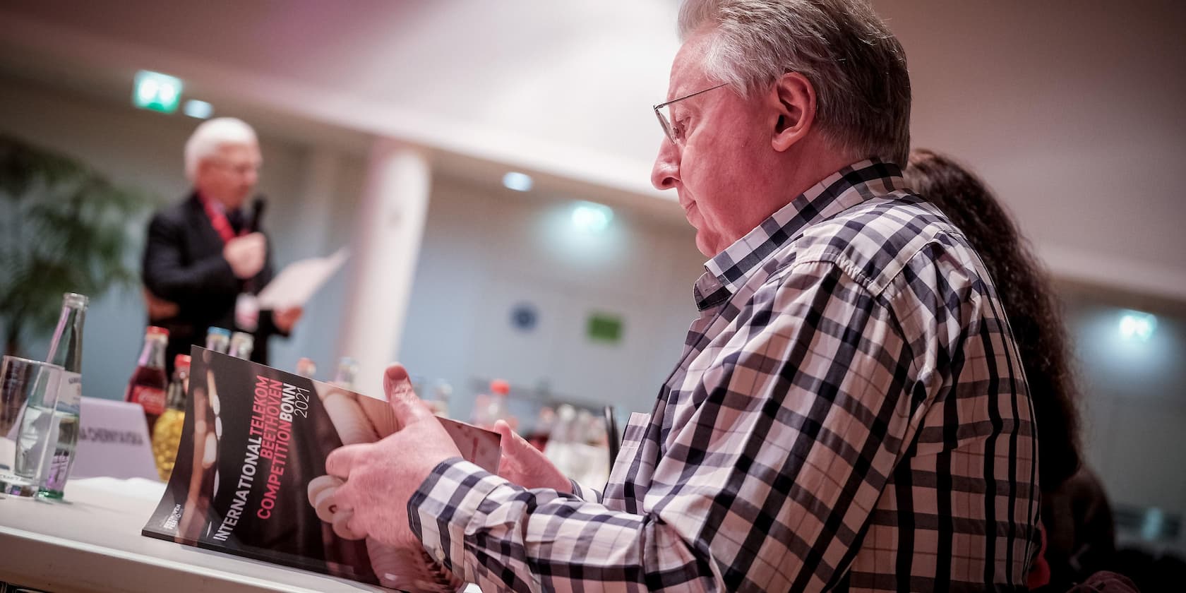 A man reads a brochure titled 'International Telekom Beethoven Competition Bonn 2021'. In the background, another man speaks into a microphone.