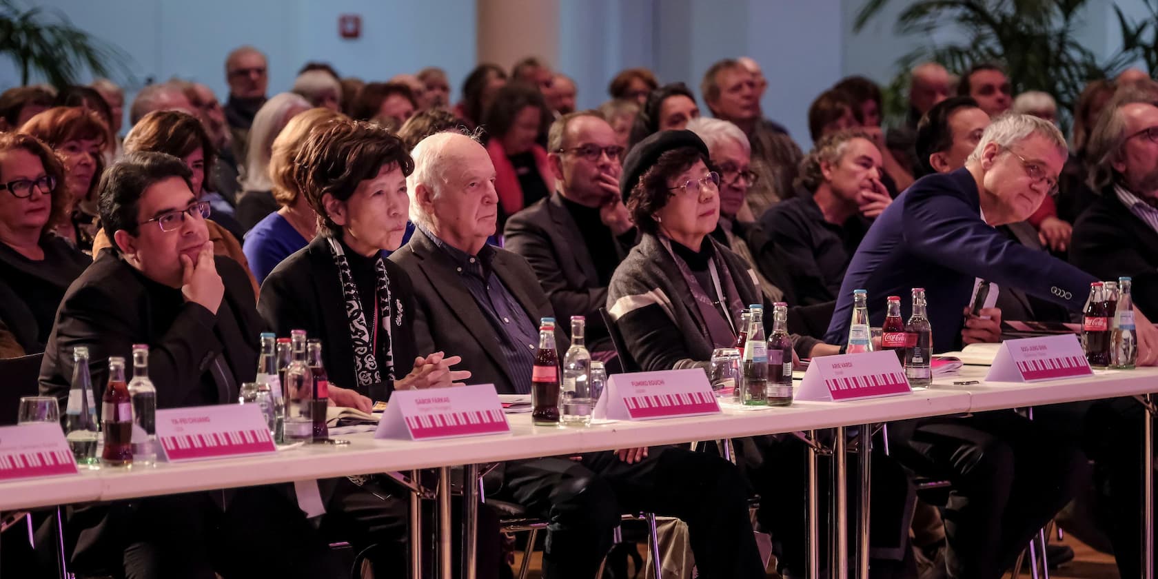 Menschen sitzen an einem langen Tisch in einem Konferenzraum und hören aufmerksam zu. Namensschilder und Getränke stehen auf dem Tisch.