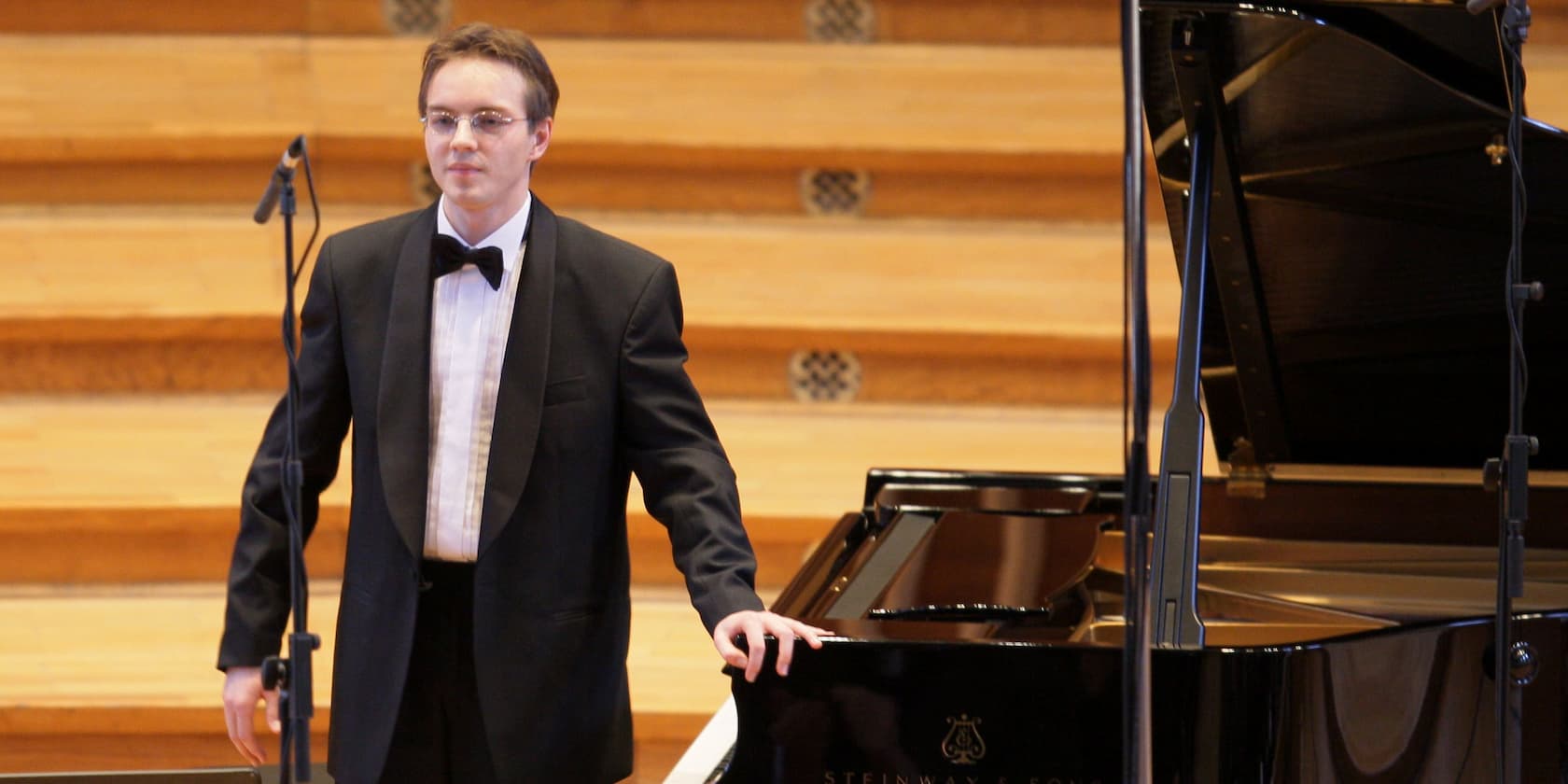 A man in a suit and bow tie stands next to a grand piano on a concert stage.