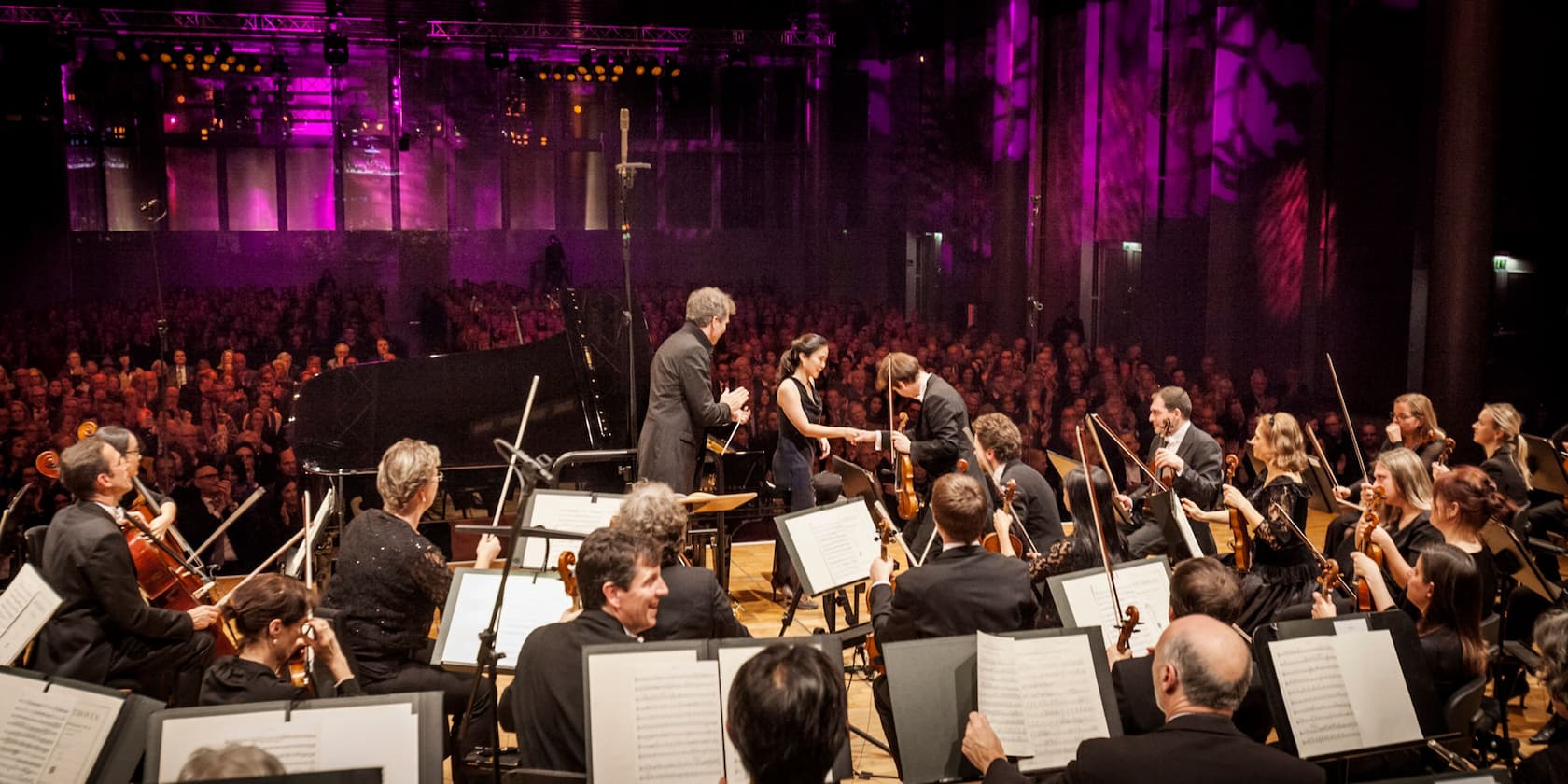 An orchestra performing in front of a large audience, with a pianist and a violinist in the foreground greeting each other. The stage is lit with purple lighting.