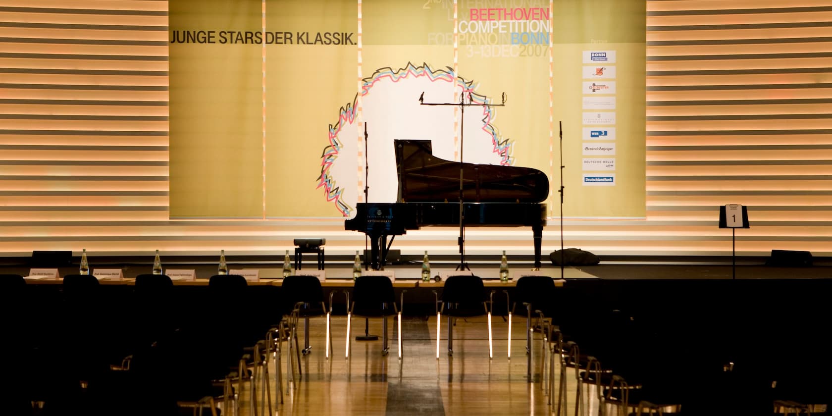 Ein Konzertsaal mit einem Flügel auf der Bühne. Im Hintergrund stehen Banner mit den Aufschriften 'Junge Stars der Klassik' und 'Beethoven Competition Bonn'.