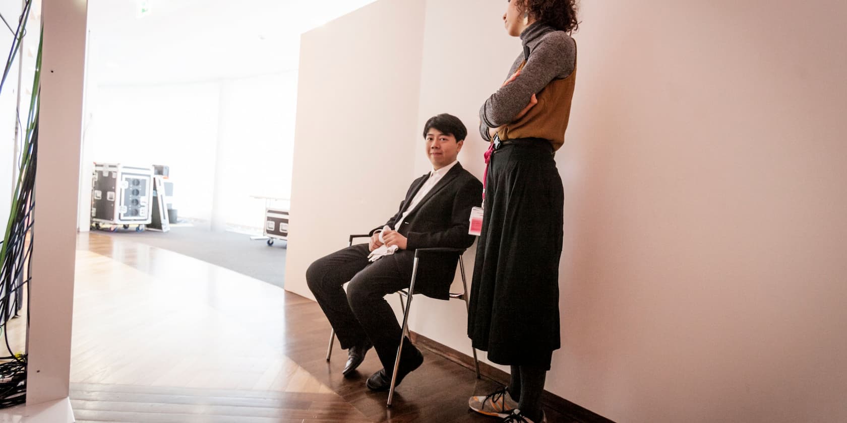 A seated person in a suit and a standing person are conversing in a bright room