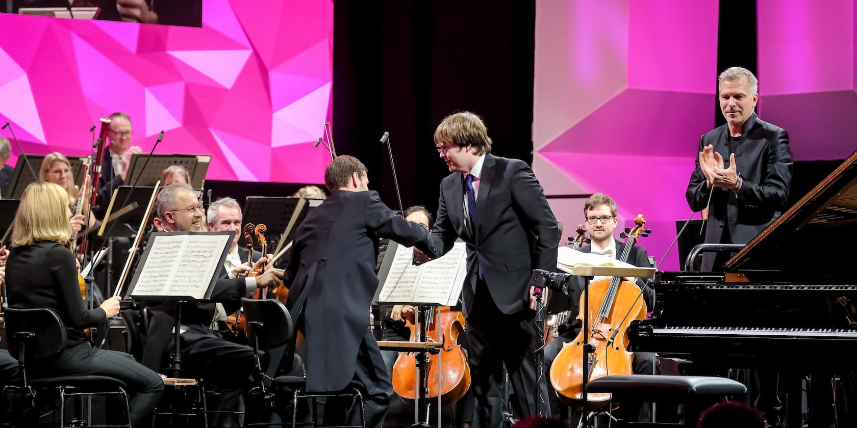 A musician shakes hands with a composer in front of an orchestra on a stage with a pink background.