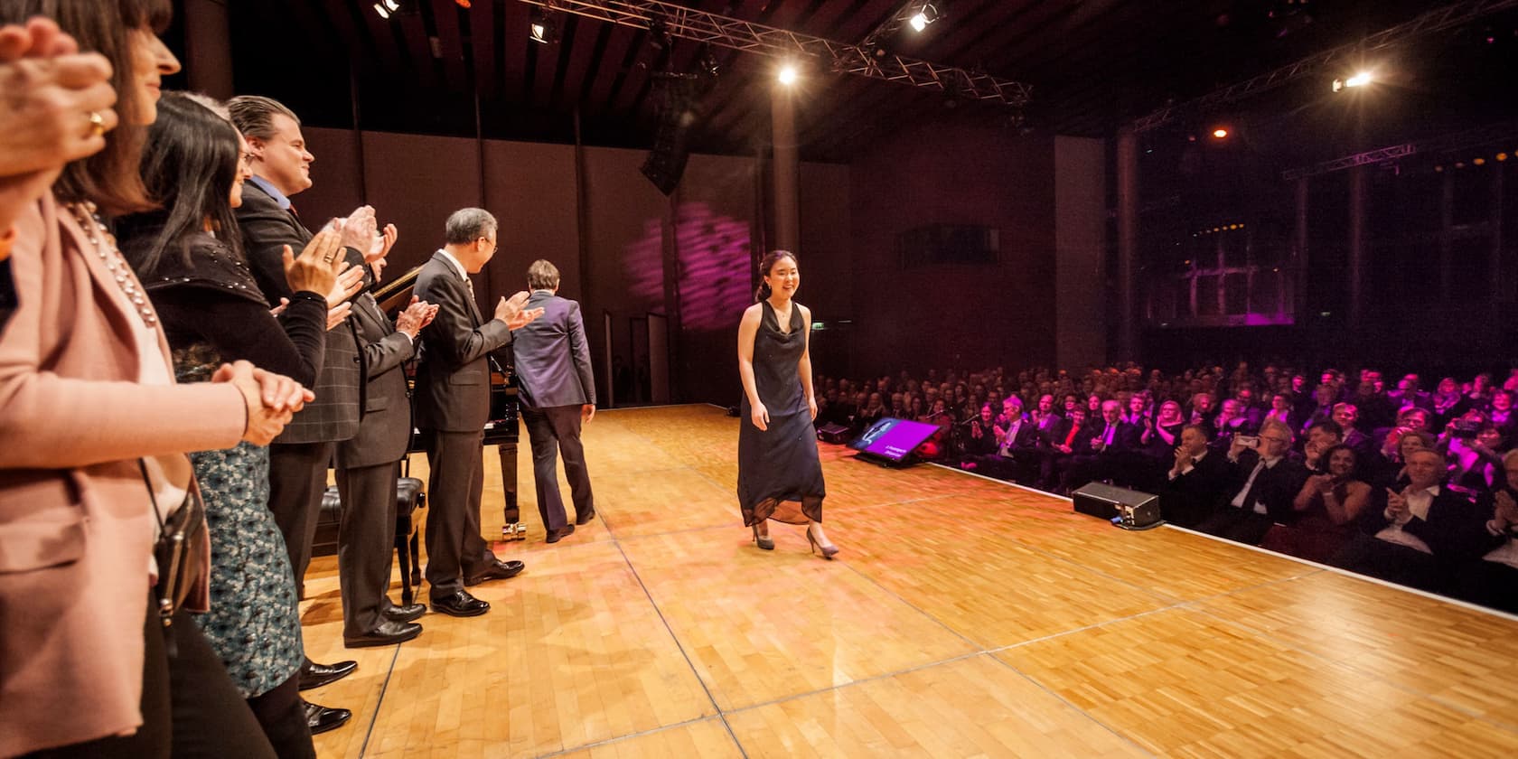 A woman in a black dress walks onto a stage while being applauded by an audience and other people on the stage.