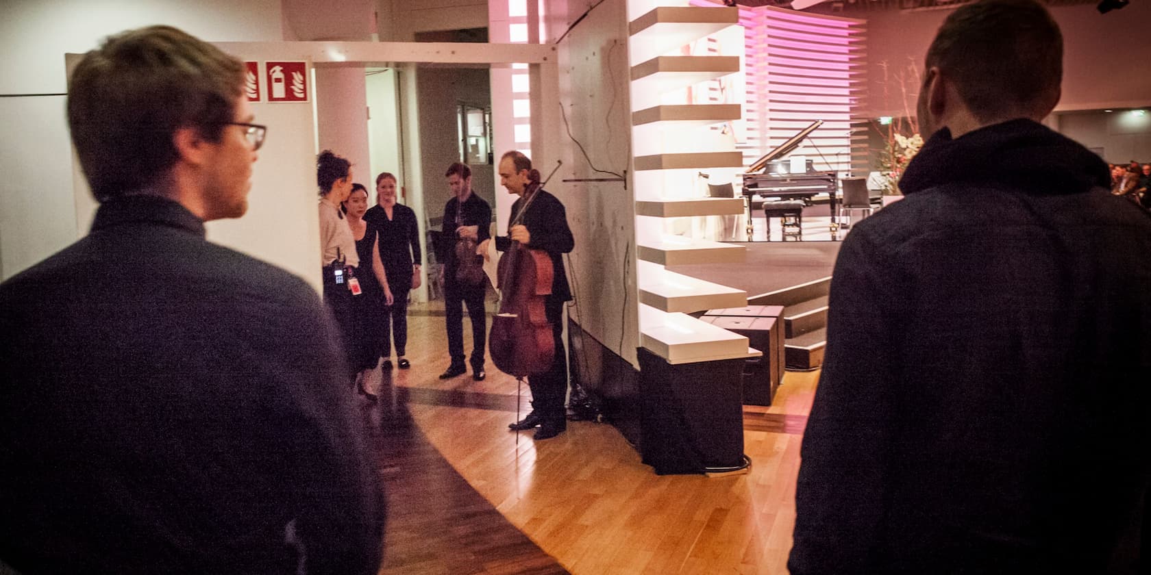 Musicians preparing backstage for a performance. A cellist and a violinist stand with their instruments, with additional people visible in the background.