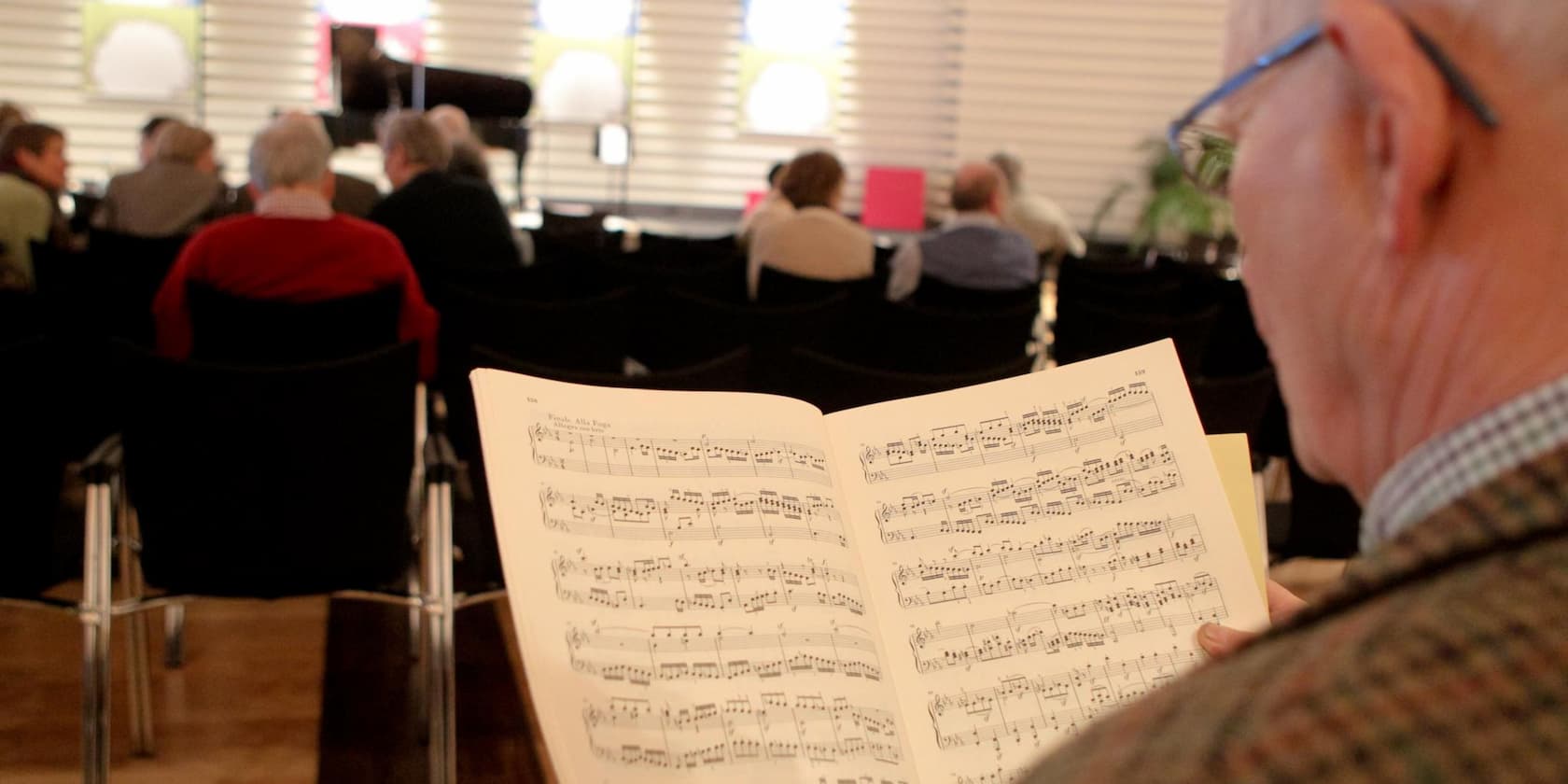 An elderly man reads sheet music in a concert hall, with other audience members in the background.