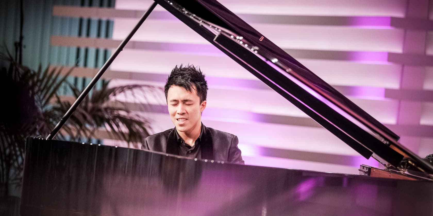 A pianist intensely plays a grand piano with closed eyes. The background is decoratively lit.