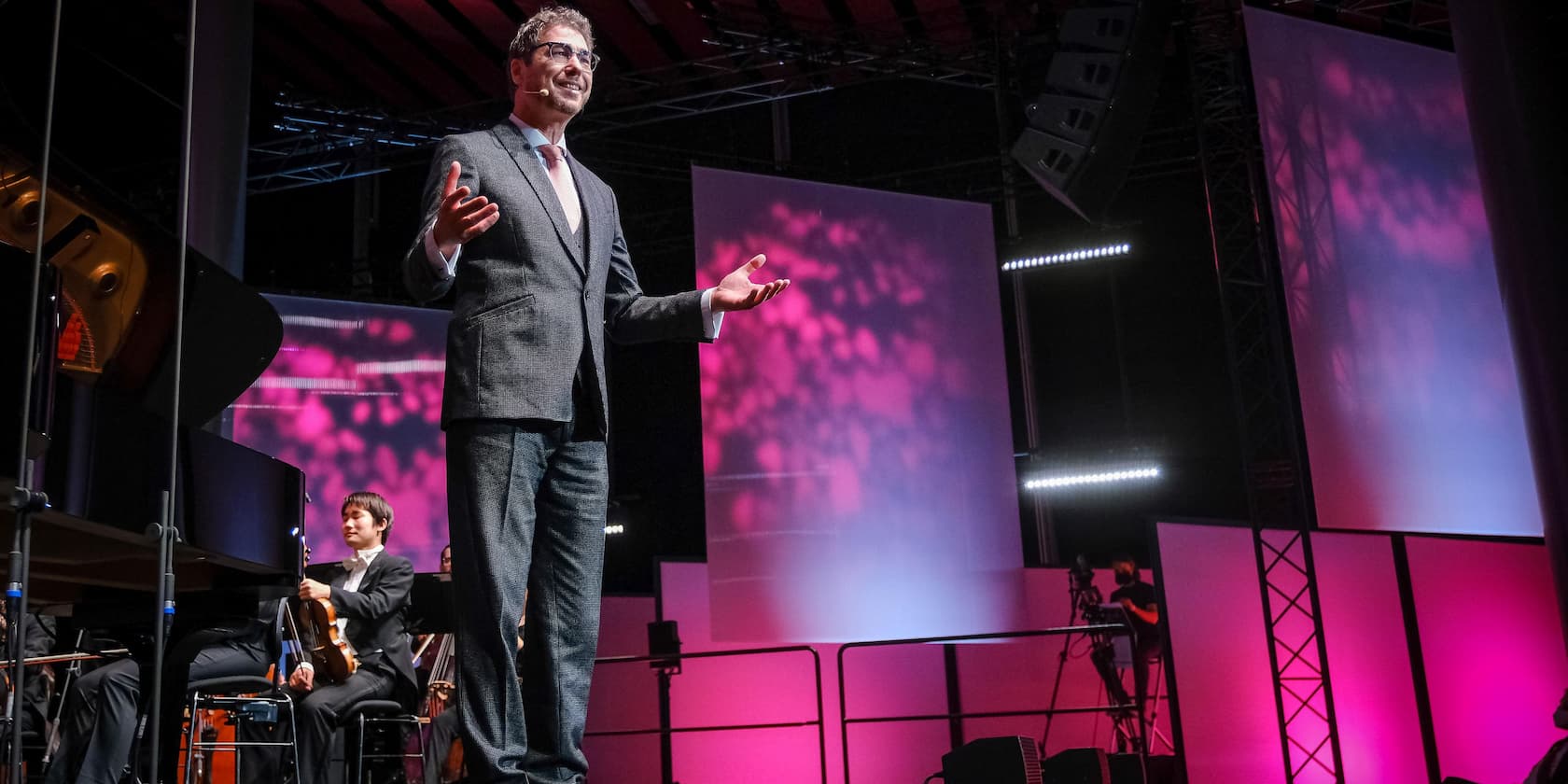 A man in a suit speaks on a stage with musicians in the background.