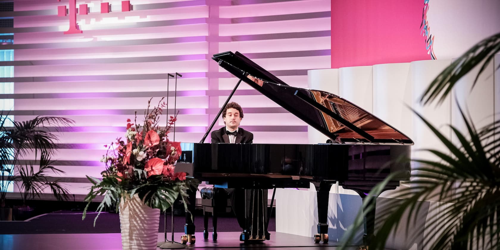 A pianist plays on a black grand piano, surrounded by plants and flowers in a modern decorated room with purple and pink lighting.