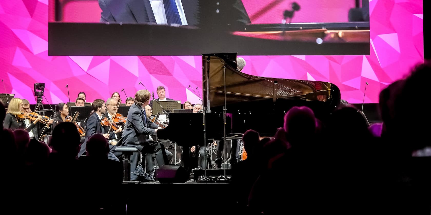 Orchestra and pianist performing on stage with a pink background, audience seated in the foreground.