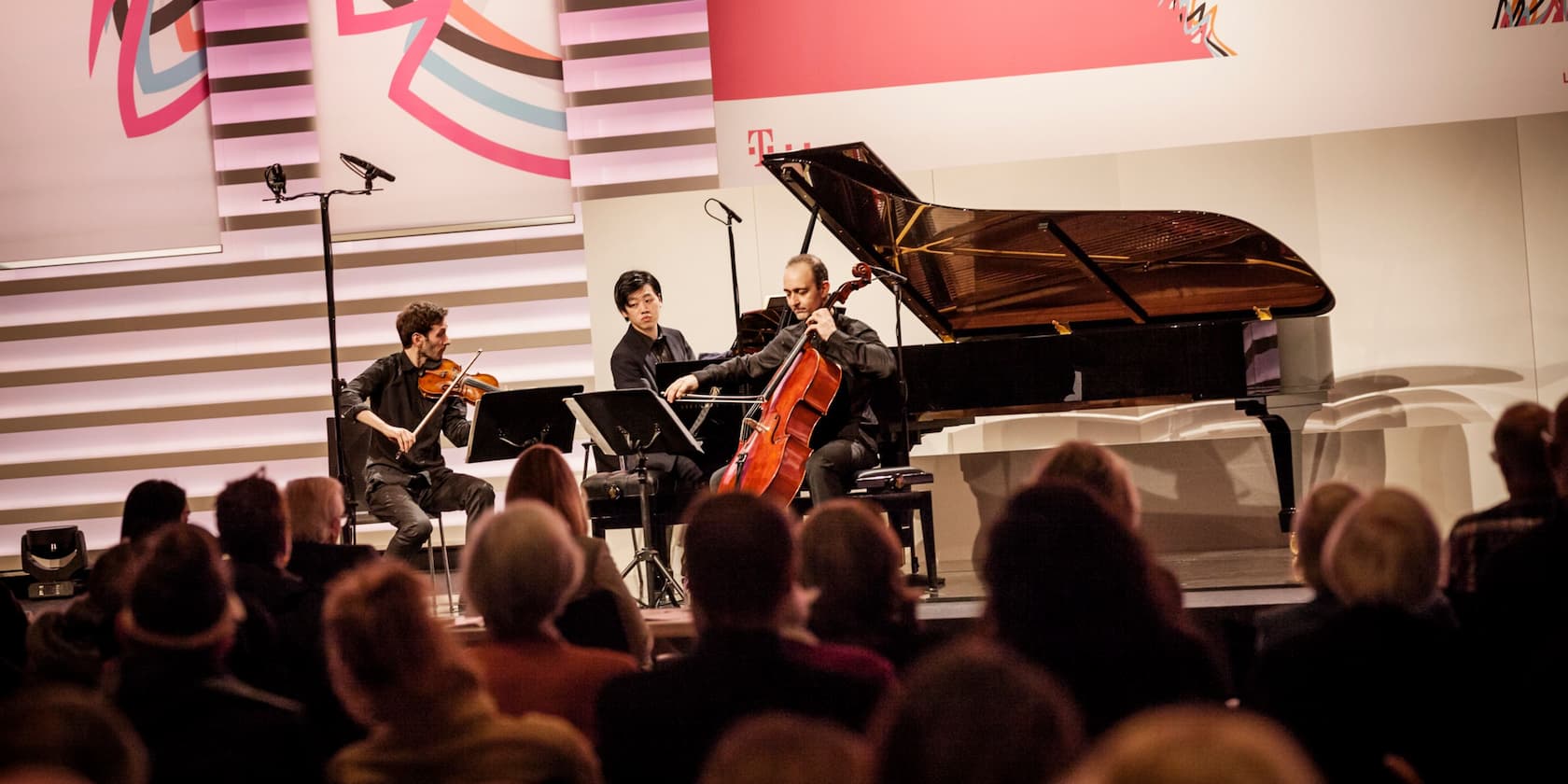 A piano trio performs on stage in front of an audience. In the background, colorful abstract designs and the Telekom logo are visible.