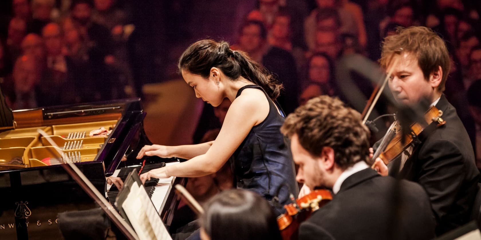 A pianist is playing intently on a piano, accompanied by two violinists on a stage.