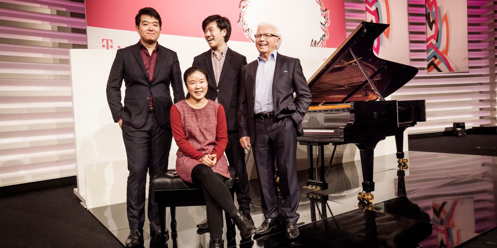 Four people stand next to a grand piano, smiling. A woman sits on a bench in front of the piano.