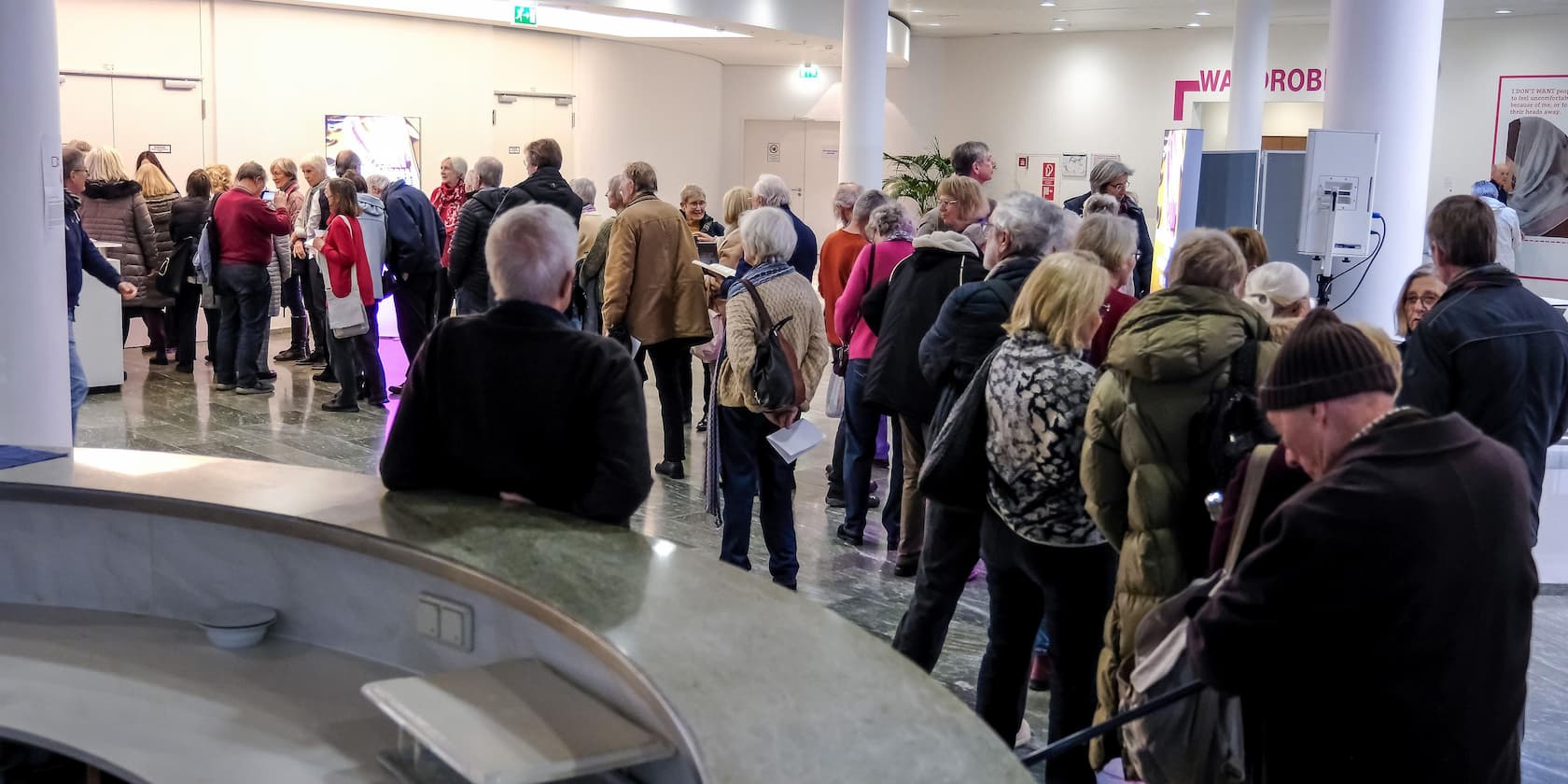 Eine Gruppe von Menschen steht in einer Schlange in einer Halle, die mit Mantelaufhänger beschriftet ist.