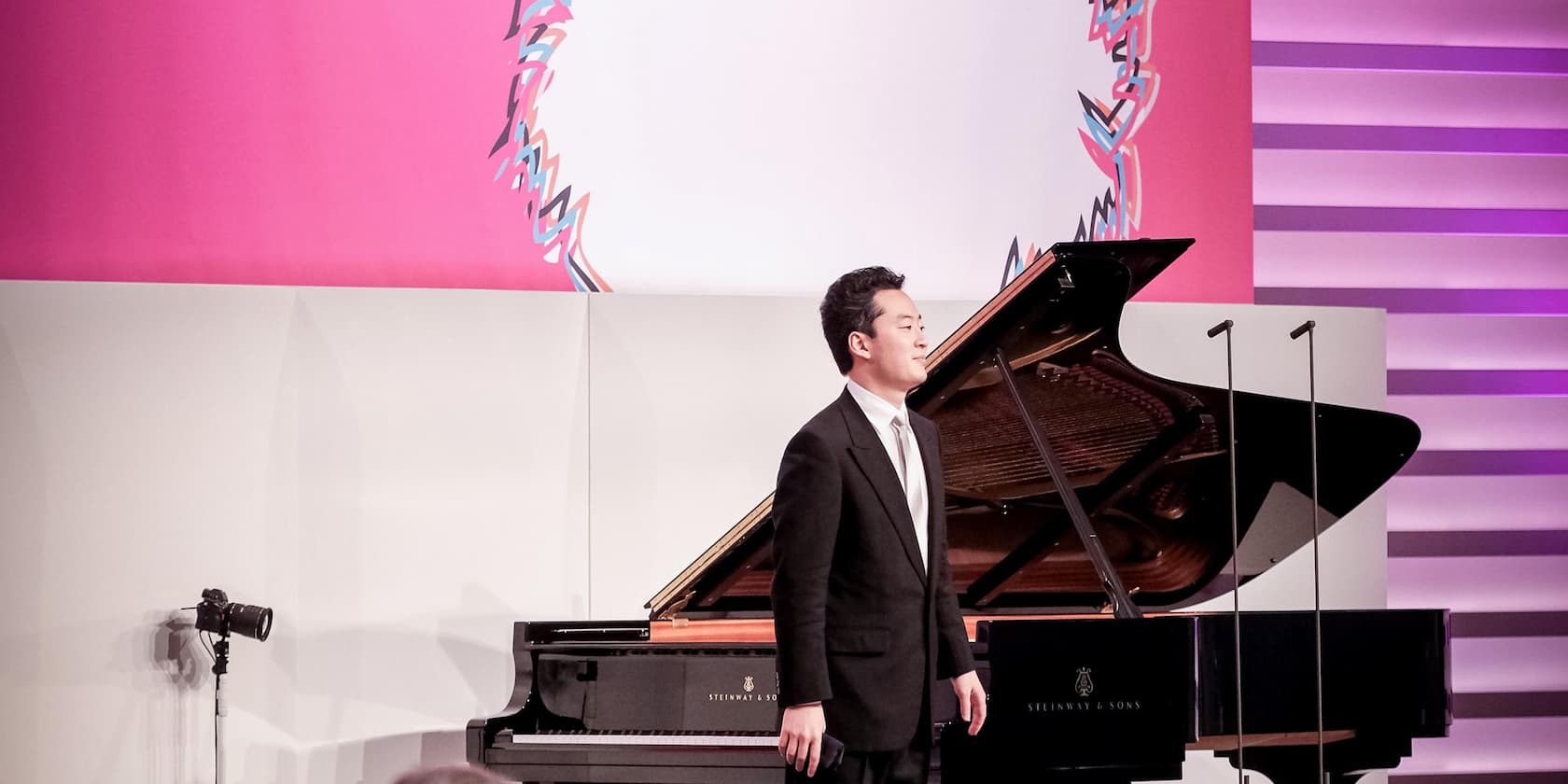 Man in a suit stands next to a grand piano on a stage with a pink background.