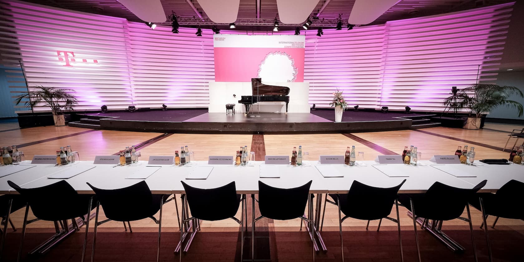 A stage with a black grand piano, behind it a purple-lit wall, and in front of the stage a table with name tags and drinks for jury members.