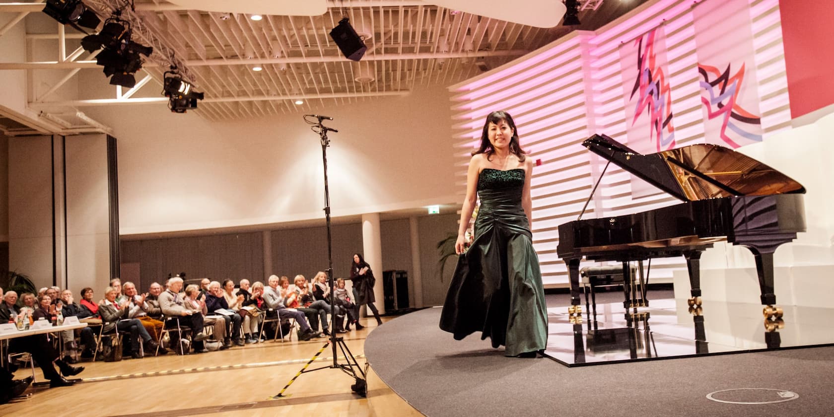 A woman in an evening dress bows after a piano concert on a stage while the audience applauds.