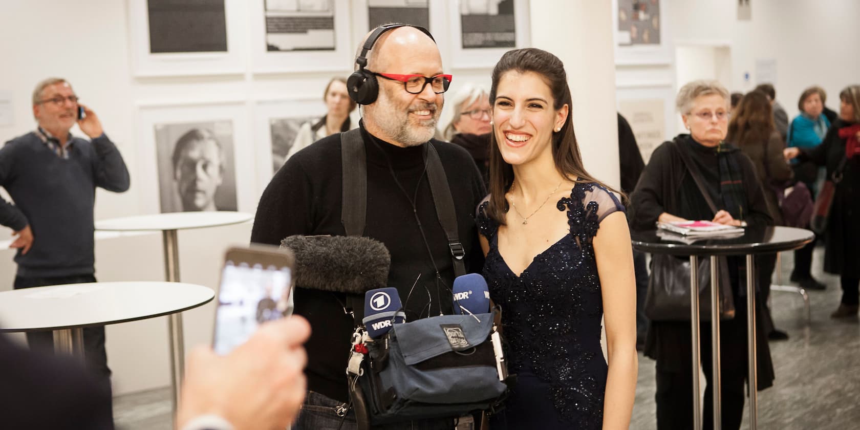 A man wearing headphones and holding a microphone with ARD and WDR logos smiles next to a woman in an evening dress. In the background, other people are standing, with some looking at the camera and talking on the phone.