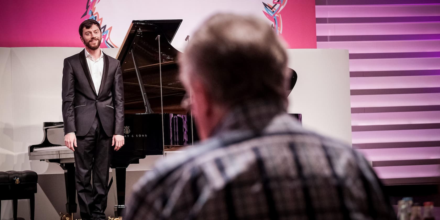 A man in a suit stands next to a grand piano on a stage, while a person sits in the foreground.