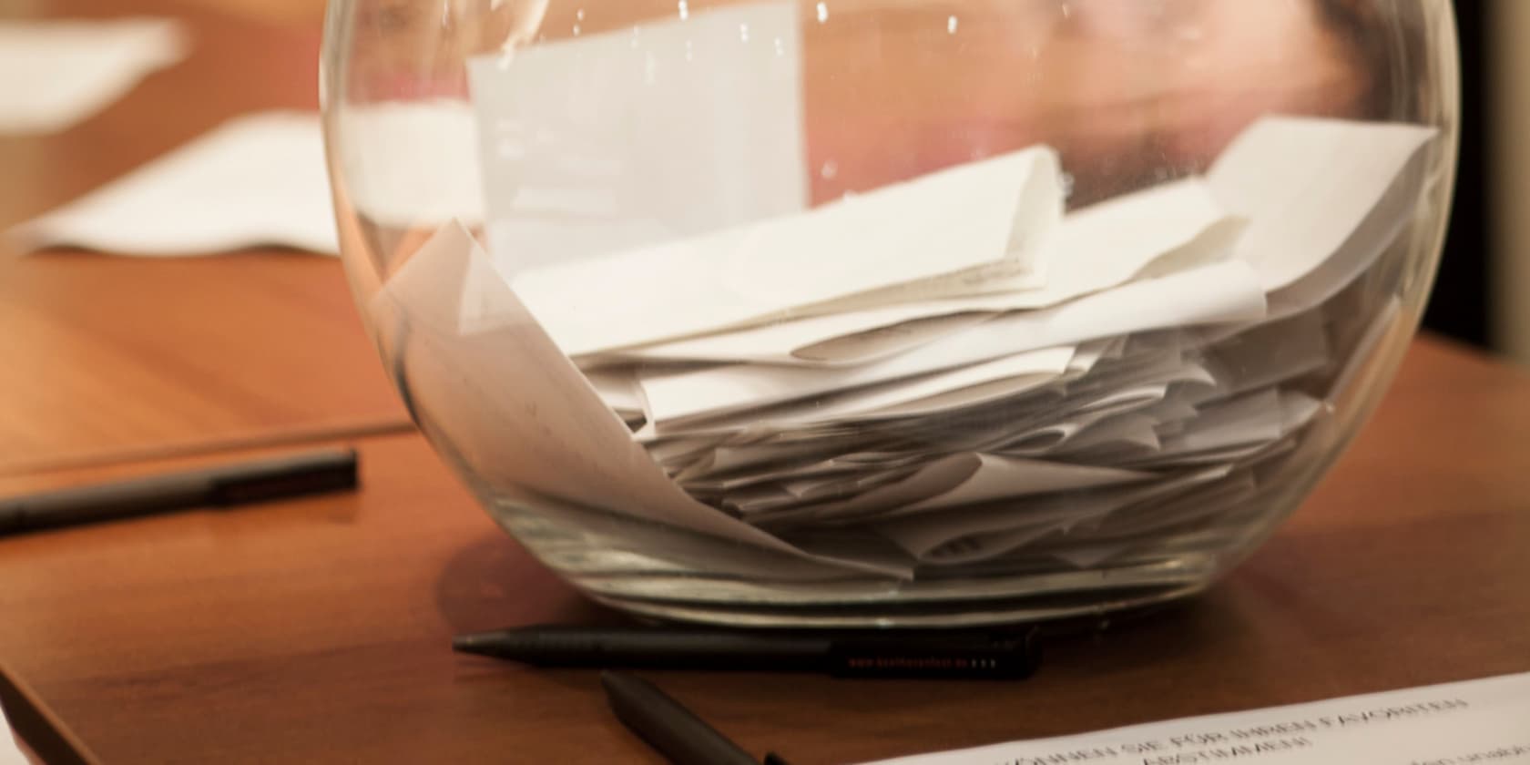 Glass bowl filled with many paper slips on a wooden table, pen in the background.