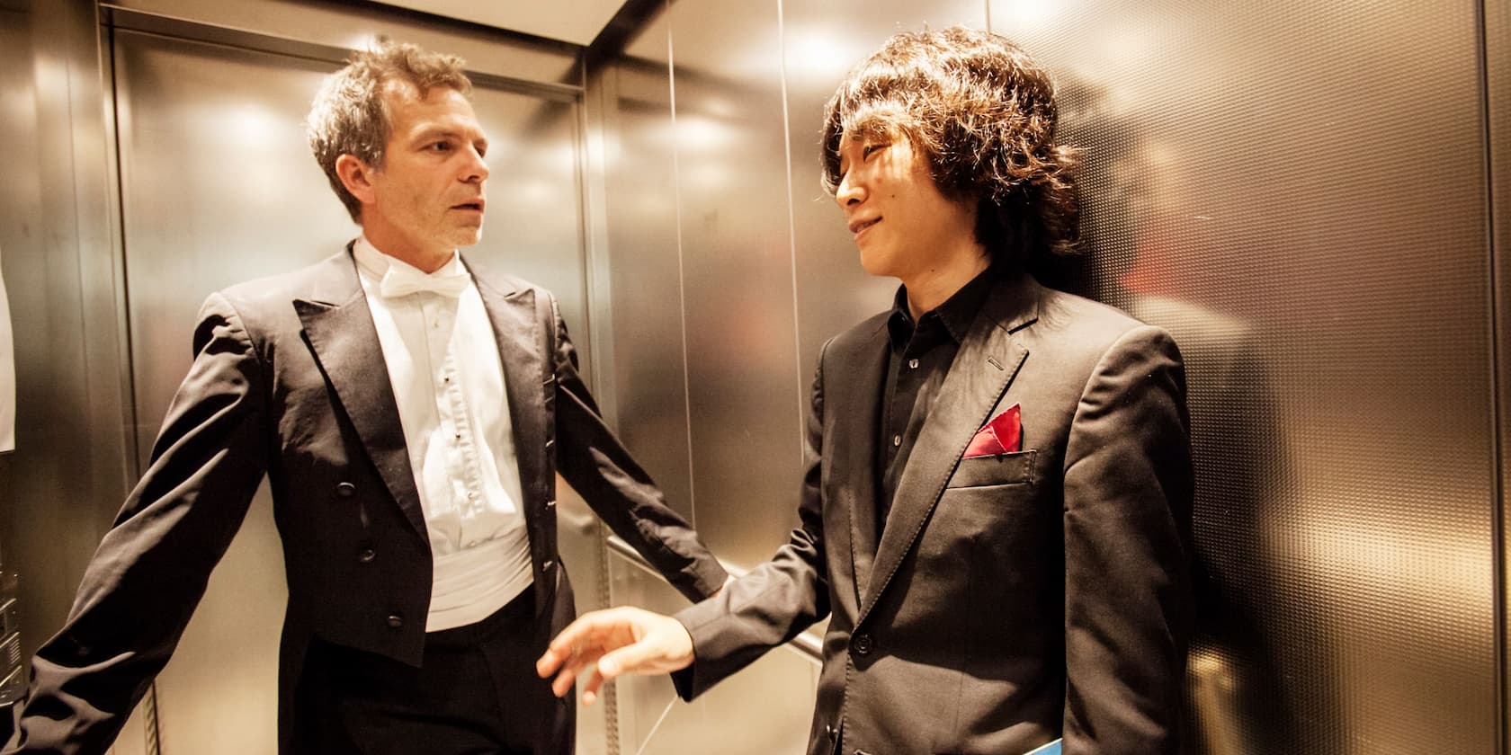 Two men in formal attire are talking in an elevator.