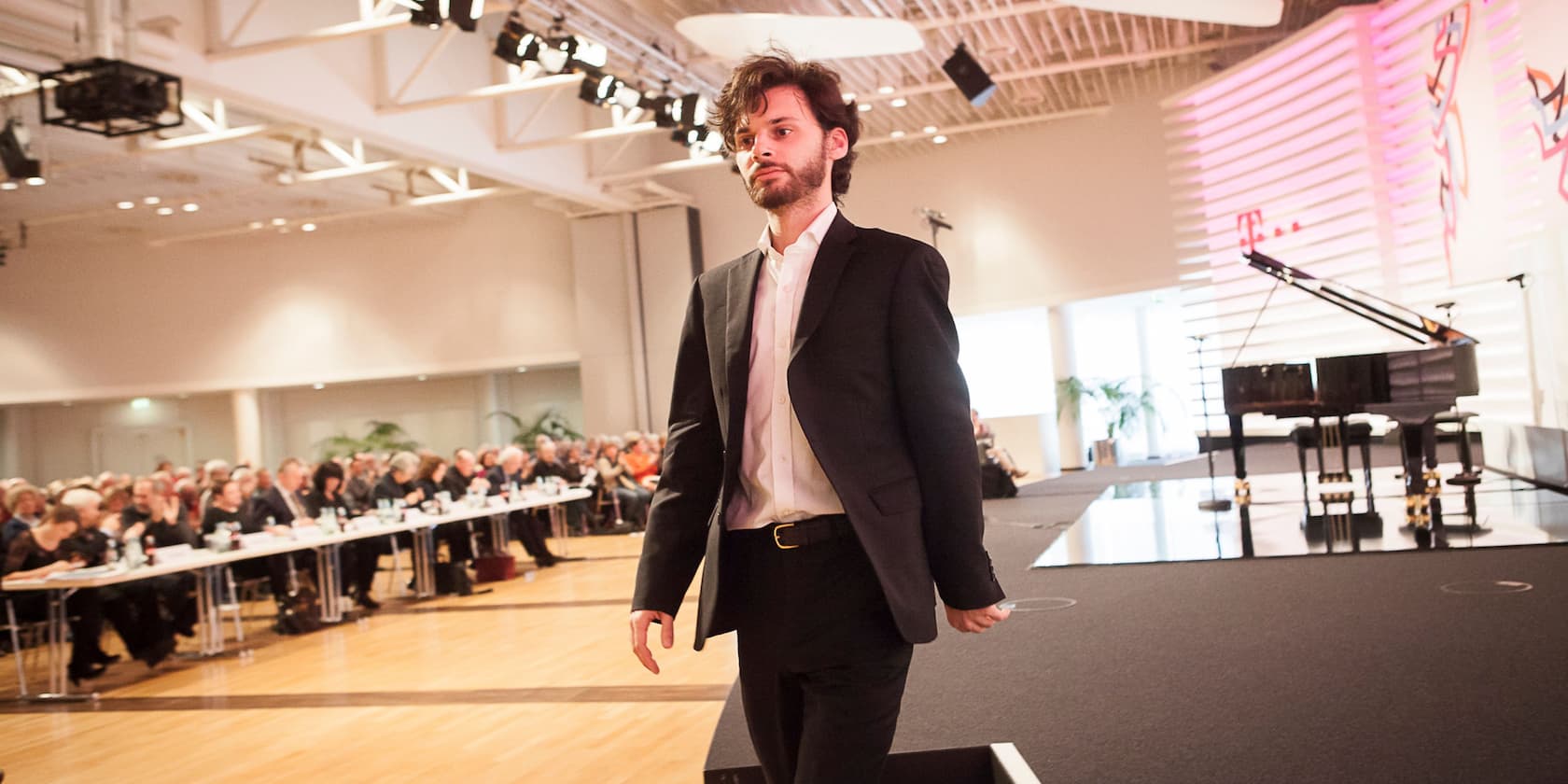 A man in a black suit stands in a conference room in front of a grand piano. In the background, many people are seated at long tables.