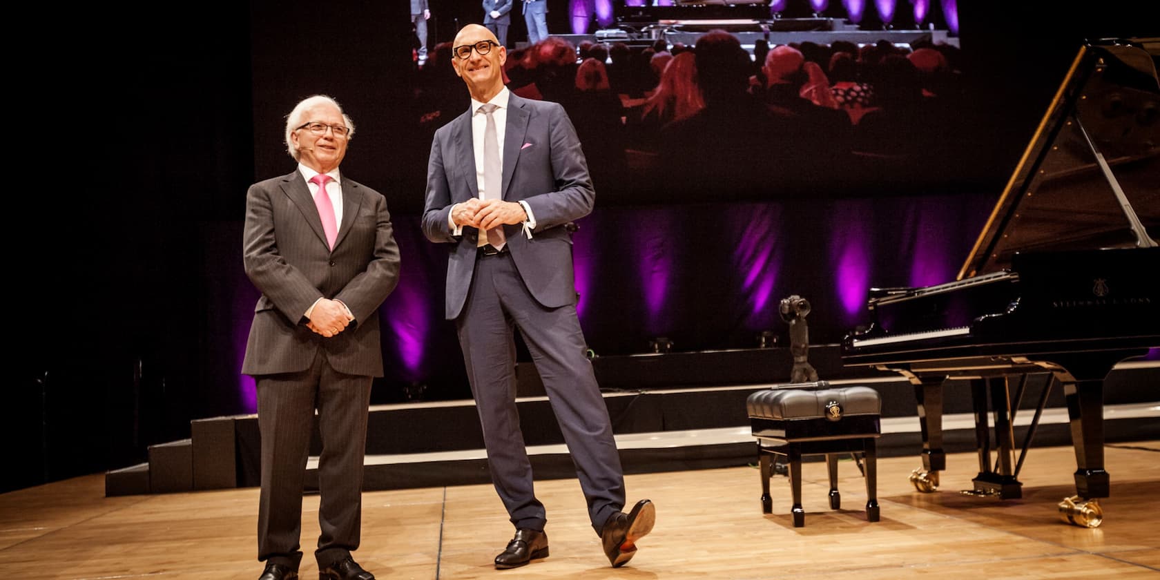 Two men in suits stand on a stage next to a Steinway grand piano; a large screen in the background shows a live broadcast.