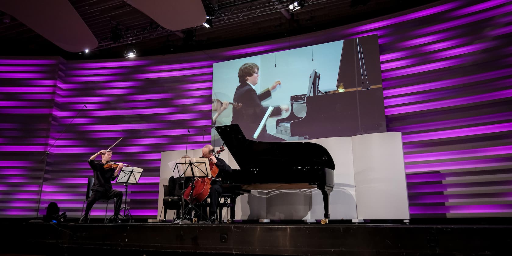 A pianist and two string musicians perform on a stage with purple backlighting. The pianist is shown on a large screen.