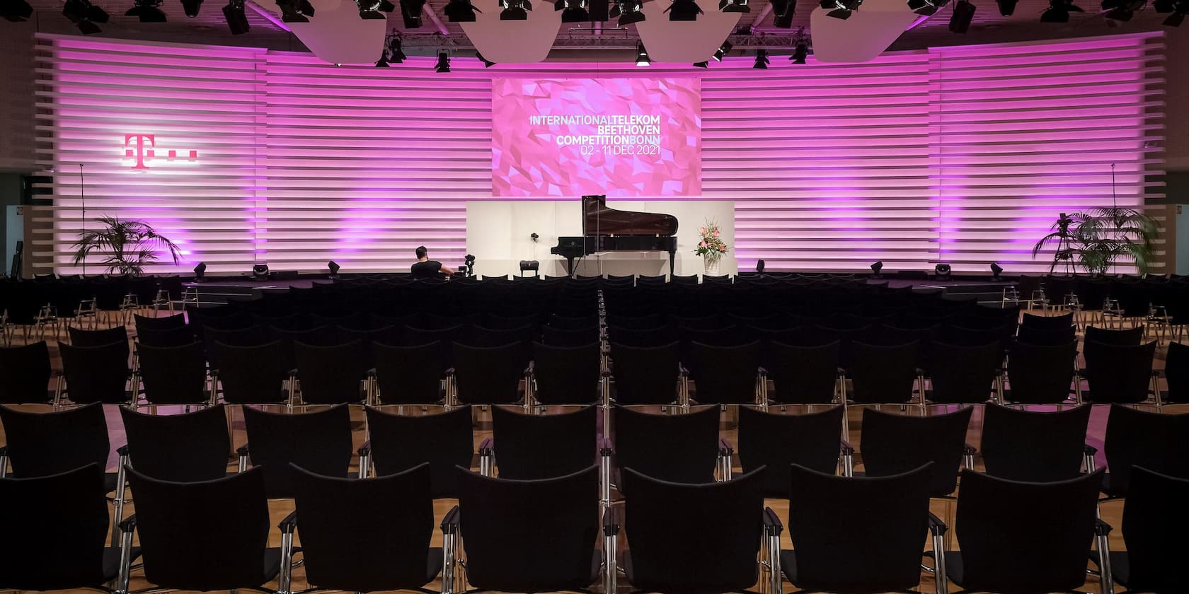 Leerer Veranstaltungssaal mit einer Bühne, die ein Klavier und blühende Pflanzen zeigt. Das Bühnenbanner zeigt den Text 'International Telekom Beethoven Competition Bonn 02 - 11 DEC 2021'.