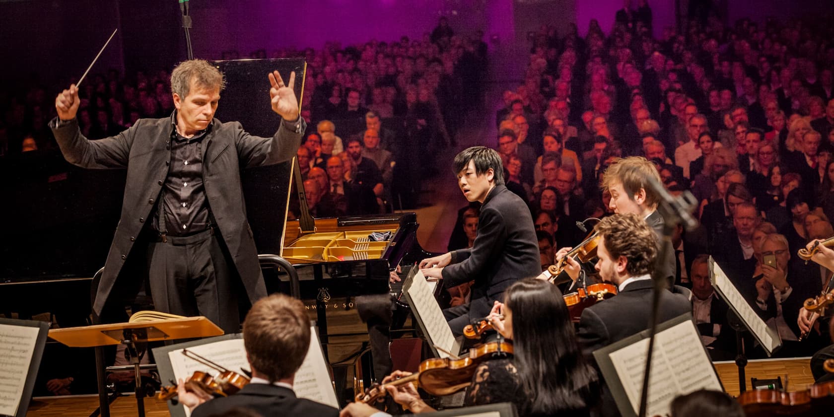 A conductor leads an orchestra with a pianist in front of an audience.