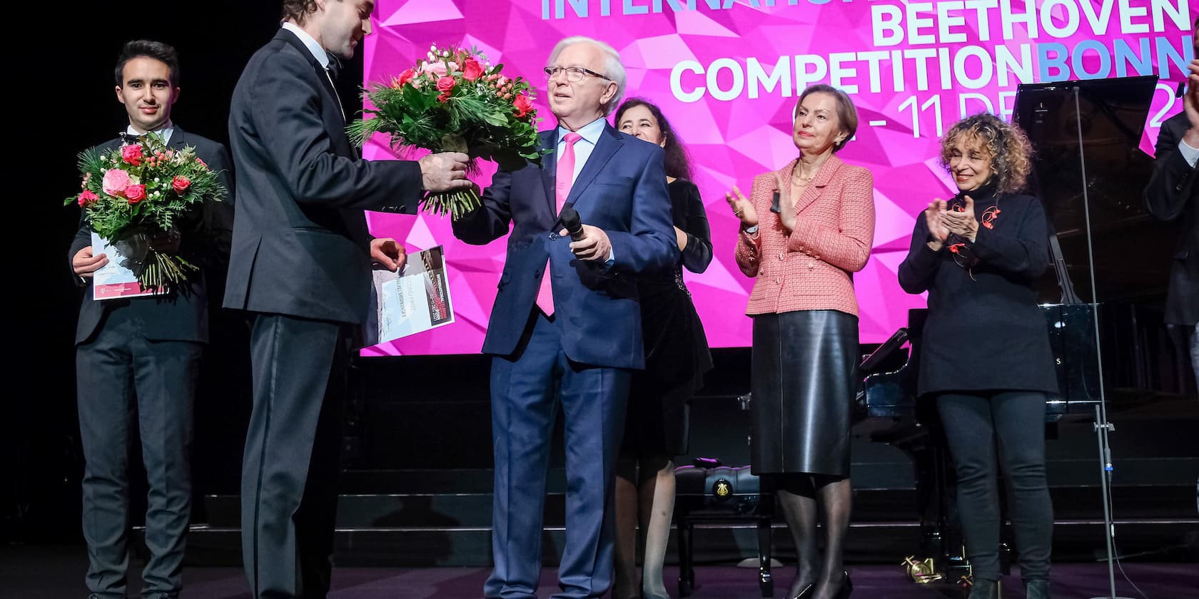 Award ceremony of the International Beethoven Competition Bonn. A man hands a bouquet of flowers to another man while other people stand nearby and applaud.