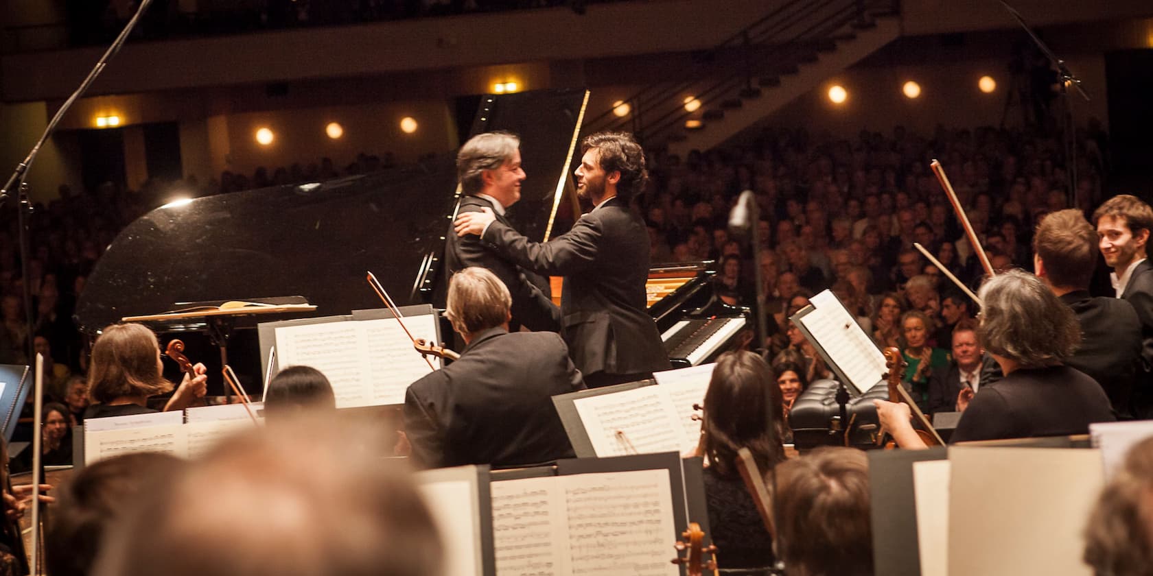 Two men in suits embrace in front of a grand piano, while the orchestra plays in the foreground and the audience applauds in the background.