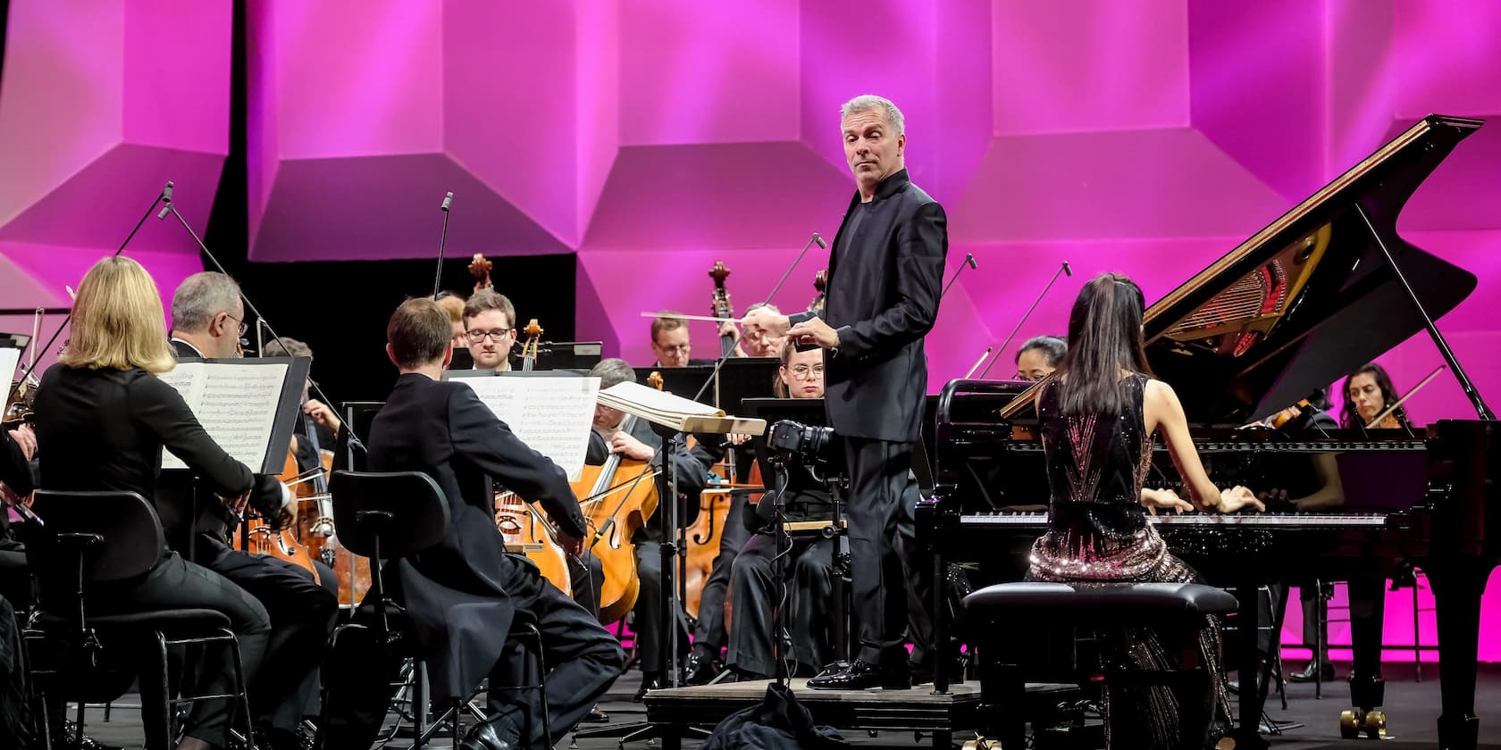 Conductor leads an orchestra during a concert with a pianist in the foreground. The background is illuminated in pink.